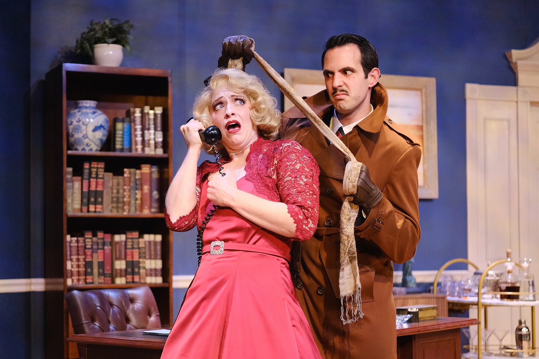 Actors Mallory Newbrough and Daniel Llaca perform a tense scene from Dial M for Murder onstage at Actors’ Playhouse, with Newbrough holding a telephone as Llaca stands behind her in a trench coat inside a 1950s-style apartment set.
