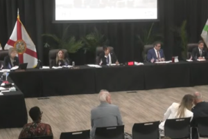 The Coral Gables City Commission seated at the dais during a public meeting, with residents visible in the foreground seating area and the Florida state flag displayed to the left.