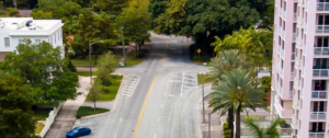 Aerial view of the Biltmore Way and Anderson Road intersection in Coral Gables. Two roadways meet at an asymmetrical junction marked with a merge lane indicator. Mature trees and a white residential structure occupy the left side of the frame; a pink high-rise building stands at right. A blue sedan is visible near the bottom left. The intersection currently operates as a two-way stop with no pedestrian crosswalks.