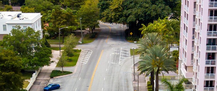 Aerial view of the Biltmore Way and Anderson Road intersection in Coral Gables. Two roadways meet at an asymmetrical junction marked with a merge lane indicator. Mature trees and a white residential structure occupy the left side of the frame; a pink high-rise building stands at right. A blue sedan is visible near the bottom left. The intersection currently operates as a two-way stop with no pedestrian crosswalks.