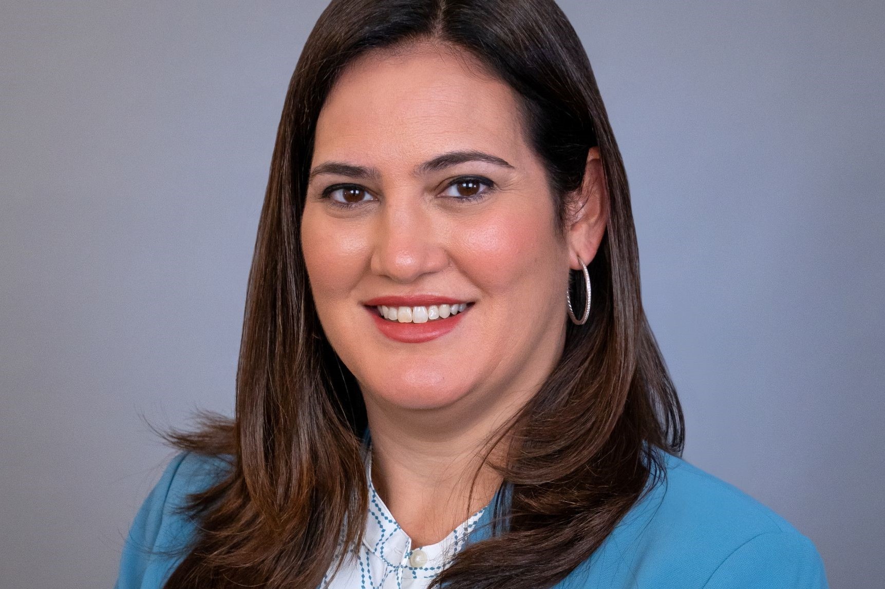 eadshot of Cristina Suárez, City Attorney of Coral Gables, wearing a teal blazer against a gray background.