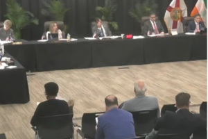 A wide shot of the Coral Gables City Commission meeting room viewed from the audience, showing five commissioners seated at a long black-draped dais with Florida and other flags visible in the background, and several members of the public seated in the foreground.