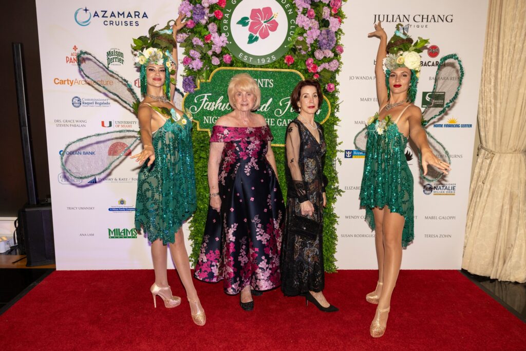 Event co-chairs Barbara Reese and Jo Wanda Peterson stand together on a red carpet in front of the Fashions & Florals sponsor backdrop, flanked by two performers in elaborate teal and green floral headdresses and sequined costumes. The backdrop features the Coral Gables Garden Club seal surrounded by flowers, with sponsor logos including Azamara Cruises, Julian Chang Boutique, Acqua Panna, Maison Perrier, and Bacardi visible on either side.