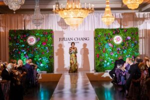 Coral Gables Garden Club President Bonnie Seipp stands on a runway in a floral gown at the Fashions & Florals gala. Behind her is a white backdrop bearing the Julian Chang Boutique logo, flanked by two lush green floral walls decorated with colorful blooms and the Coral Gables Garden Club seal. Crystal chandeliers hang overhead and formally dressed guests are seated at tables on either side of the runway.