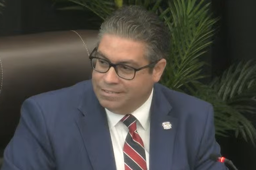 Coral Gables Commissioner Ariel Fernandez seated at the dais during a City Commission meeting, wearing a blue suit, white shirt and striped tie.