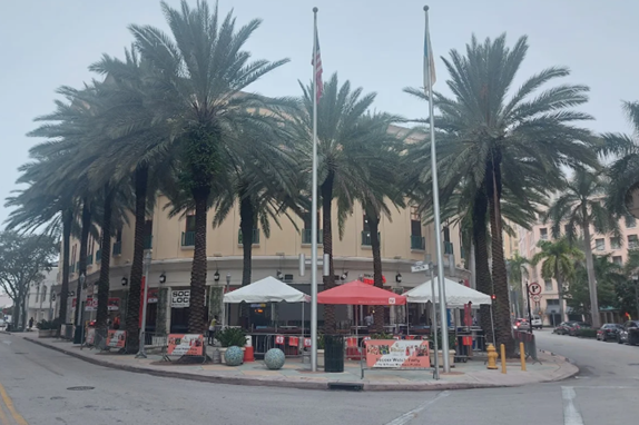The exterior of Fritz and Franz Bierhaus at the corner of Merrick Way in Coral Gables, showing the restaurant's outdoor plaza with white and red umbrellas, palm trees, and two flagpoles flying the American and another flag. The Mediterranean-style building behind the plaza is visible through the tree canopy.