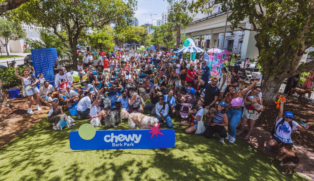 A large crowd of pet owners and their dogs pose for a group photograph at Chewy Bark Park in Coral Gables. The crowd fills the frame from foreground to background along The Underline's tree-lined corridor. A blue Chewy Bark Park sign is prominently displayed in the foreground. Colorful balloons, pool floats, and event installations are visible throughout. Attendees are cheering, waving, and holding their dogs.
