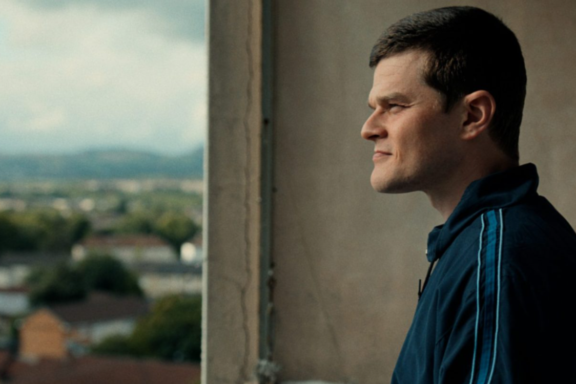 A man stands in profile by a window, looking out over a townscape, in a scene from the film I swear.