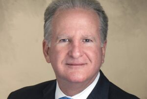 Professional headshot of Peter J. Iglesias, Coral Gables City Manager. A man with gray-streaked hair in a dark suit, white shirt, and blue tie faces the camera with a neutral expression against a beige background.