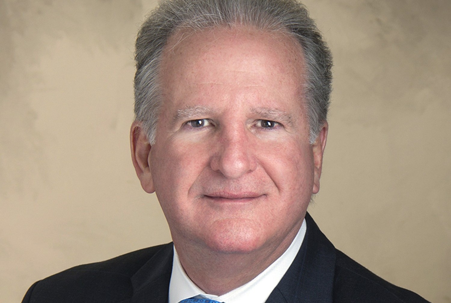 Professional headshot of Peter J. Iglesias, Coral Gables City Manager. A man with gray-streaked hair in a dark suit, white shirt, and blue tie faces the camera with a neutral expression against a beige background.