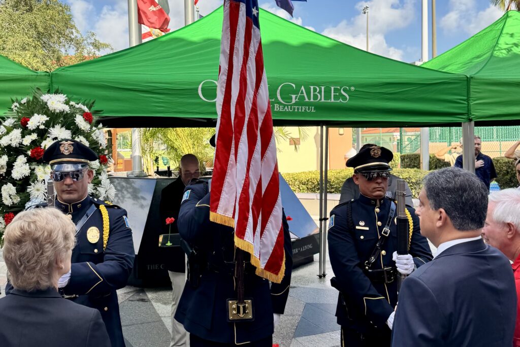 Members of the Coral Gables Honor Guard present the American flag during the city’s Memorial Day ceremony at the War Memorial Youth Center, with attendees gathered around the memorial monument beneath green City of Coral Gables tents.