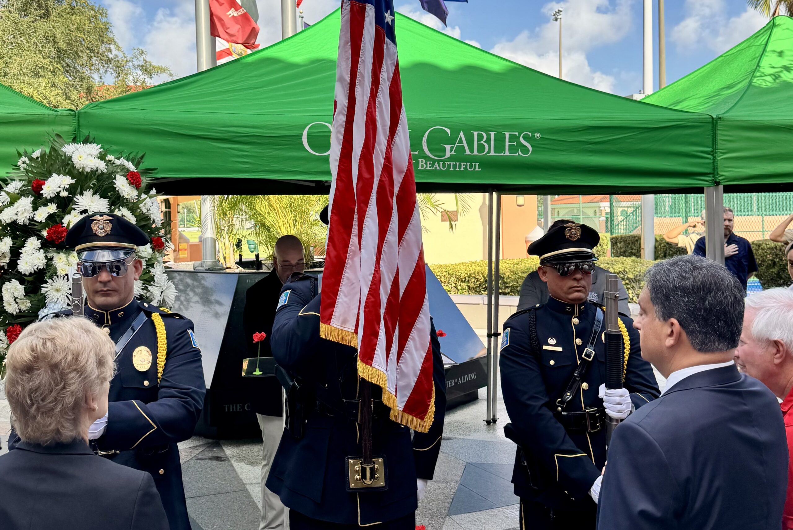 Members of the Coral Gables Honor Guard present the American flag during the city’s Memorial Day ceremony at the War Memorial Youth Center, with attendees gathered around the memorial monument beneath green City of Coral Gables tents.