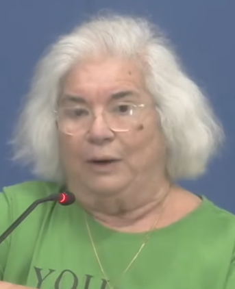 Coral Gables resident Maria Cruz speaking at a public comment podium, an older woman with white hair and glasses wearing a green shirt, a microphone visible in front of her.