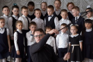 A young man in a dark shirt and glasses conducts with a baton in the foreground while approximately two dozen young children in formal school uniforms — white shirts, dark vests, and dark dresses — stand arranged in rows behind him against a draped white backdrop. The children's expressions range from focused to uncertain.