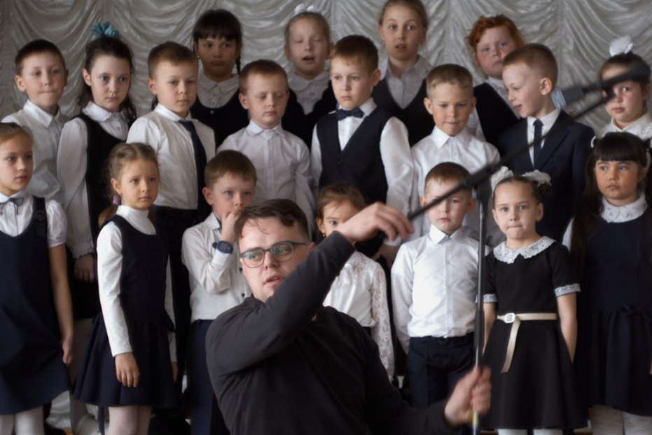 A young man in a dark shirt and glasses conducts with a baton in the foreground while approximately two dozen young children in formal school uniforms — white shirts, dark vests, and dark dresses — stand arranged in rows behind him against a draped white backdrop. The children's expressions range from focused to uncertain.