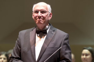 Robert Longfield stands on a concert stage in a dark tuxedo and bow tie, holding a conductor's baton at his side. He faces the camera with a composed expression. Musicians and a warmly lit stage backdrop are visible behind him.