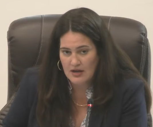 Coral Gables City Attorney Cristina Suarez speaking at the commission meeting, seated at the dais in a navy blazer, with dark hair and hoop earrings, a microphone visible in front of her.