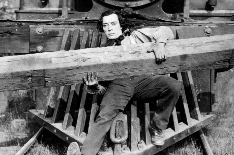 A black and white still from the 1926 silent film The General, showing Buster Keaton in period clothing lying across the cow catcher at the front of a steam locomotive, his expression characteristically deadpan as he balances on the wooden frame with the engine behind him.