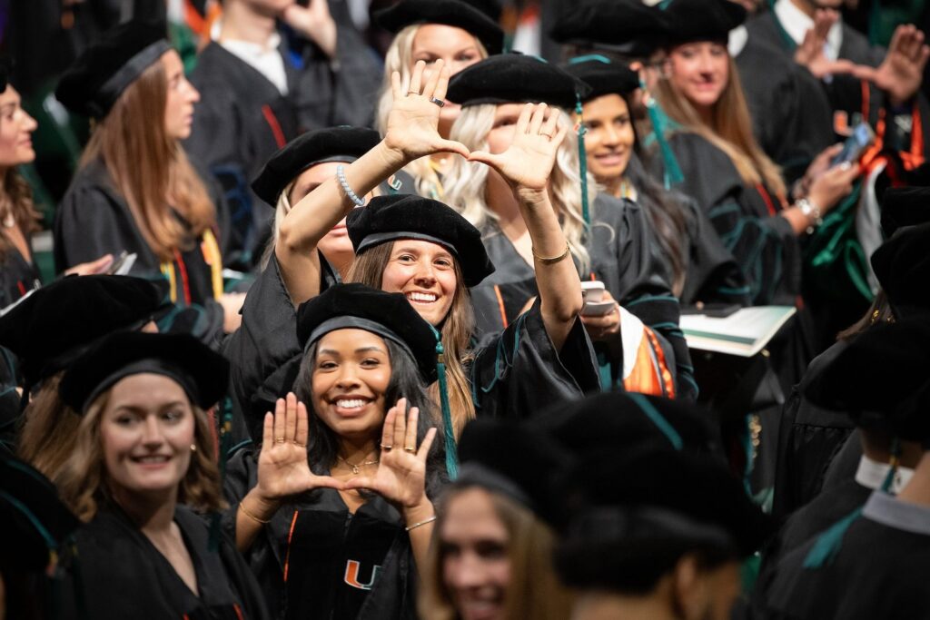 A crowd of graduates in black doctoral gowns and velvet tam caps at the University of Miami commencement. Two women in the foreground smile and hold their hands up forming the U gesture, the university's signature hand sign. Orange and green hood trim is visible on several graduates' regalia.