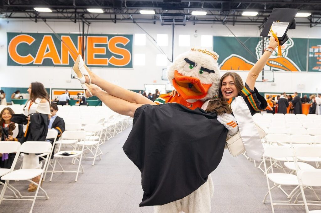 A smiling female graduate in a black gown is lifted off the ground by Sebastian the Ibis, the University of Miami mascot, inside the Watsco Center. She holds her diploma cover aloft with one hand and raises her legs in the air. A large green and orange "Canes" banner is visible on the wall behind them.