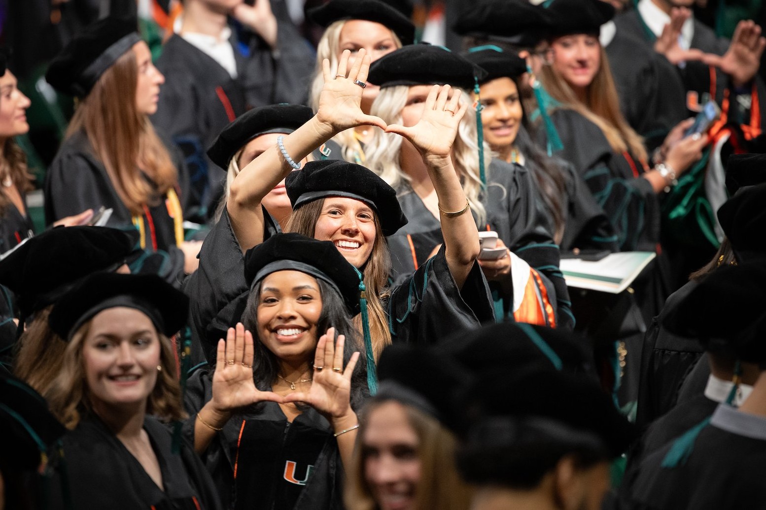 A crowd of graduates in black doctoral gowns and velvet tam caps at the University of Miami commencement. Two women in the foreground smile and hold their hands up forming the U gesture, the university's signature hand sign. Orange and green hood trim is visible on several graduates' regalia.