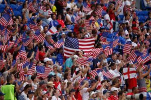 A packed stadium section filled with fans waving American flags and English St. George's Cross flags, many dressed in red, white, and blue, cheering during an international soccer match.