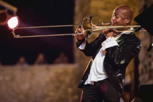 Cuban trombonist Julio Montalvo performs onstage at night, playing trombone under warm concert lighting during an outdoor live music performance.