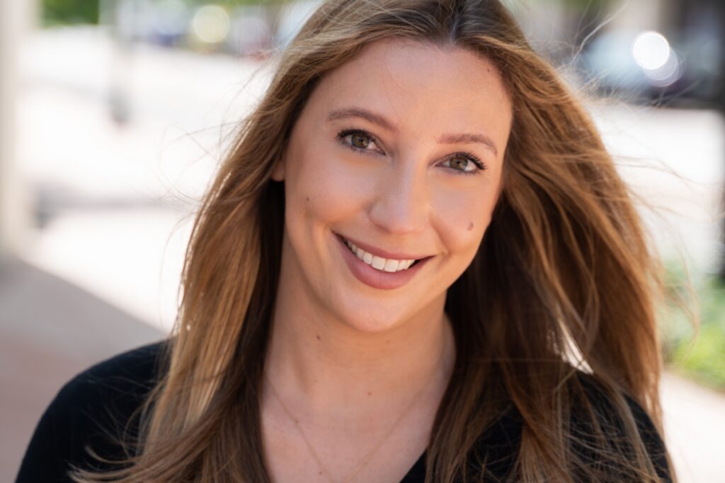 Head-and-shoulders portrait of Lindsay Kerdyk smiling outdoors, with long light-brown hair and a softly blurred background.