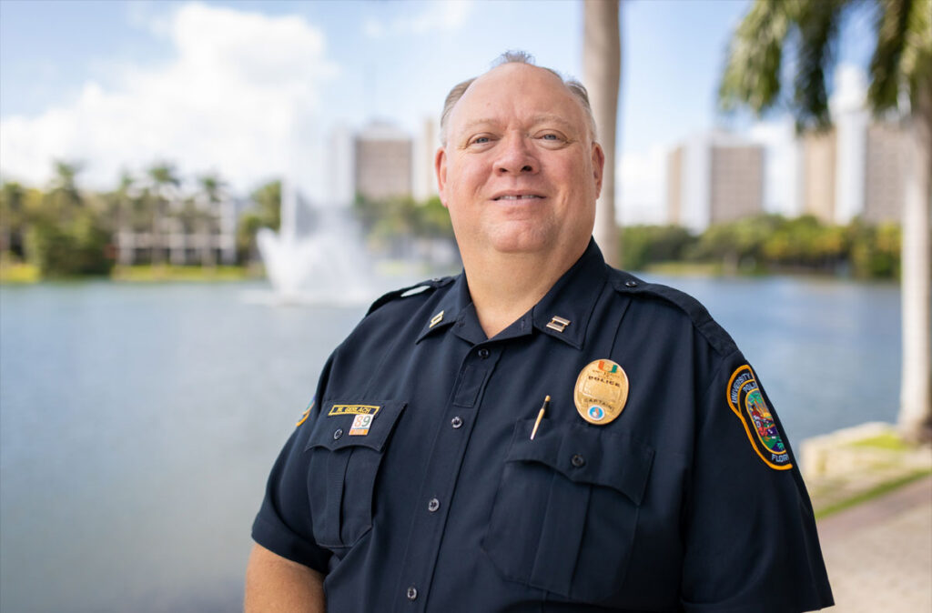 Portrait of University of Miami Police Captain William Gerlach in uniform, standing outdoors beside a campus lake with palm trees, a fountain, and university buildings in the background.