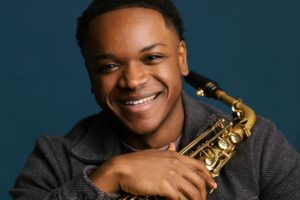 Smiling saxophonist holding a gold saxophone against a dark blue background.