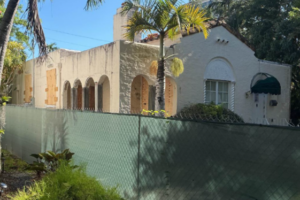 Cream-colored building behind a green construction fence, with boarded openings, arched windows, palm trees and overgrown landscaping.