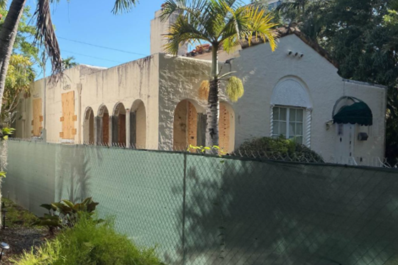 Cream-colored building behind a green construction fence, with boarded openings, arched windows, palm trees and overgrown landscaping.