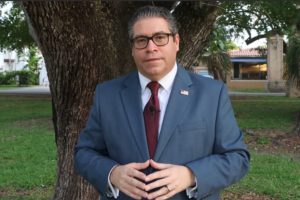 Coral Gables Commissioner Ariel Fernandez stands outdoors in a suit and tie in front of a large tree.