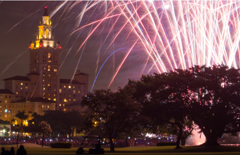 Fireworks explode over the Biltmore Hotel at night, with trees and spectators visible in the foreground.