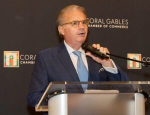 Man in a blue suit speaks into a handheld microphone at a podium in front of a Coral Gables Chamber of Commerce backdrop.