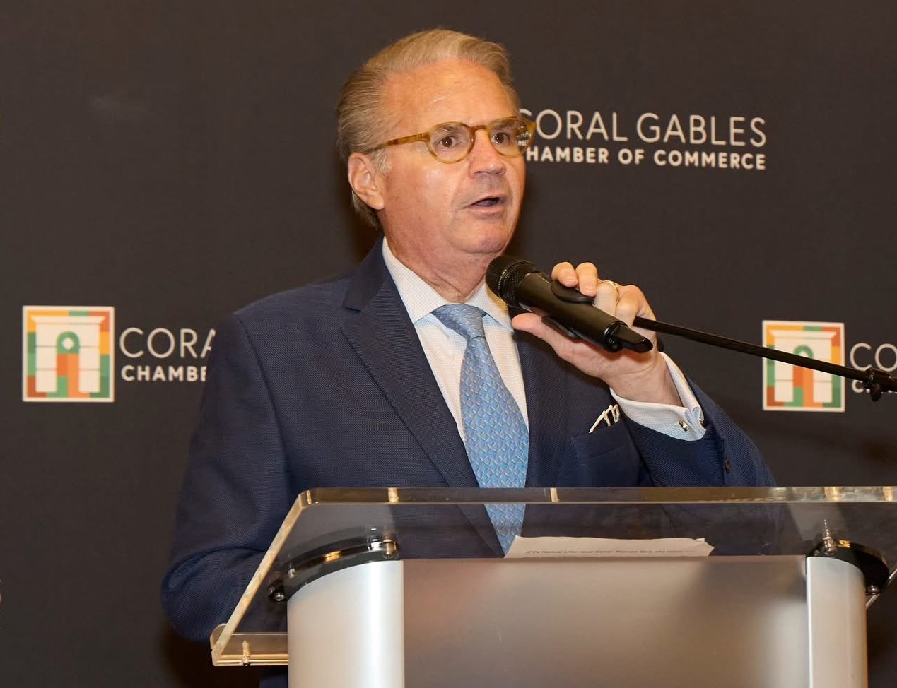 Man in a blue suit speaks into a handheld microphone at a podium in front of a Coral Gables Chamber of Commerce backdrop.