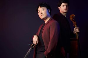 Cellists Ken Kubota and Mitchell Lyon pose back to back with their instruments against a dark studio background.