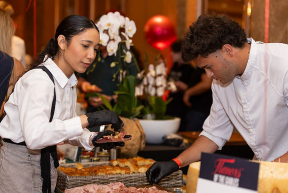 Two servers in white shirts and black gloves prepare and serve food at a restaurant tasting station during an indoor event.