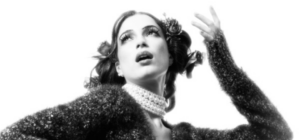 Black-and-white promotional portrait of Luzía looking upward with one hand raised, wearing floral hair adornments and a pearl choker.