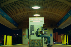 A young man sits alone on a concrete bench at night beneath the arched roof of a transit station.