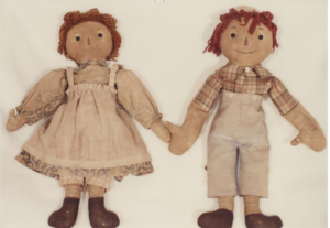 Two vintage Raggedy Ann and Andy cloth dolls lie side by side against a light background, holding hands.
