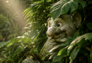 Stone troll figure with large eyes and pointed ears partially hidden among dense green tropical plants.