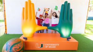 Three fans pose behind oversized orange and green University of Miami hands forming the split U at a FIFA Fan Festival booth.