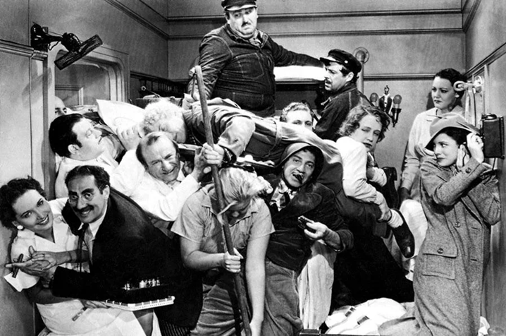 Black-and-white film still showing a crowded ship stateroom packed with people, luggage and performers in a comic pileup.