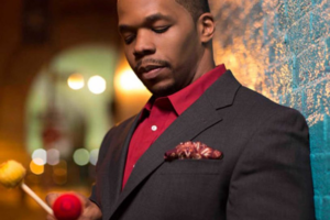 Warren Wolf, wearing a dark jacket and red shirt, holds vibraphone mallets while standing against a colorful background.
