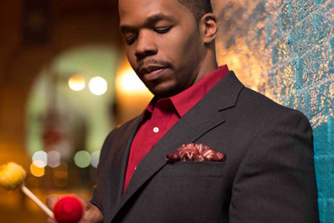 Warren Wolf, wearing a dark jacket and red shirt, holds vibraphone mallets while standing against a colorful background.