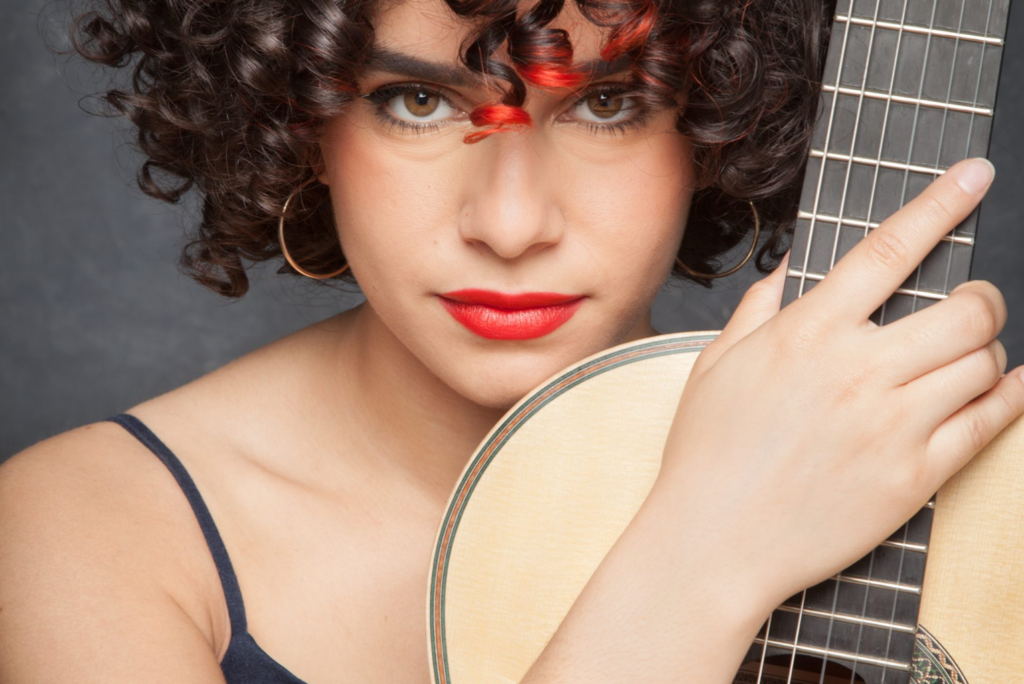 Close-up portrait of a woman holding a classical guitar, with curly dark hair, red lipstick and a guitar neck beside her face.