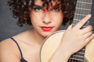 Close-up portrait of a woman holding a classical guitar, with curly dark hair, red lipstick and a guitar neck beside her face.