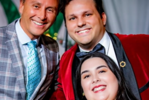 Two smiling men in formal jackets pose behind a smiling woman in a close-up promotional photograph.