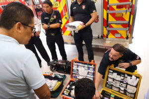 Coral Gables firefighters display open cases of emergency medical supplies to a group of visiting professionals beside a fire-rescue vehicle.
